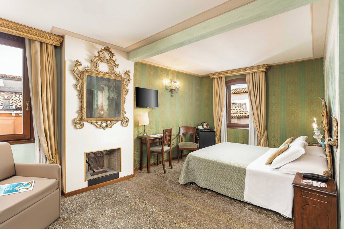 Habitación de hotel con cama, escritorio con silla, chimenea, televisor y ventana con cortinas.