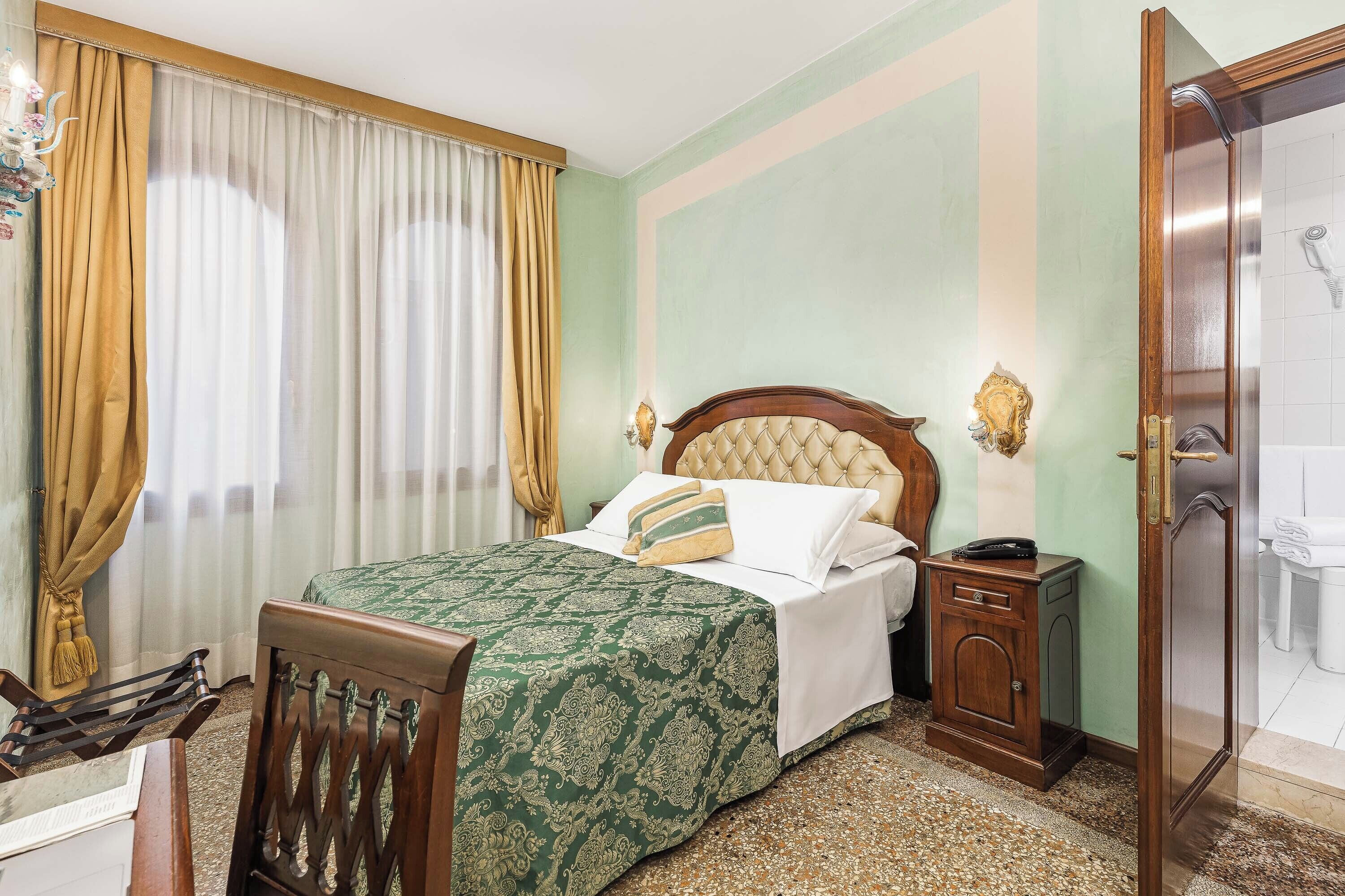 A neatly arranged hotel room with a bed, a wooden nightstand, a chair, a bathroom with a shower, and a window with curtains.