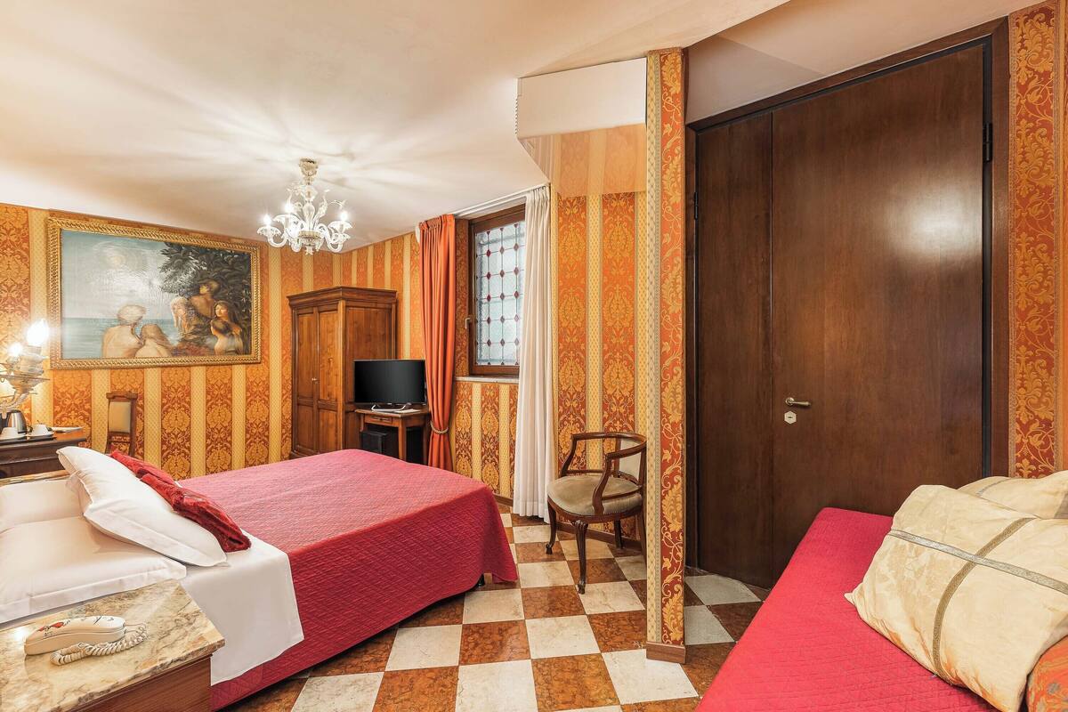 Una habitación de hotel con piso a cuadros, una cama con colcha roja, un armario de madera, un televisor y un cuadro en la pared.