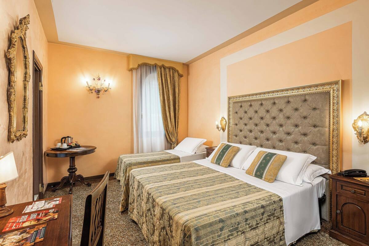 Habitación de hotel con dos camas, una mesa redonda con lámpara, una mesita con hervidor y un candelabro.