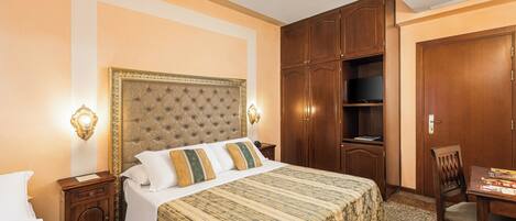 A hotel room with a large bed, wooden wardrobe, and a desk with a chair.