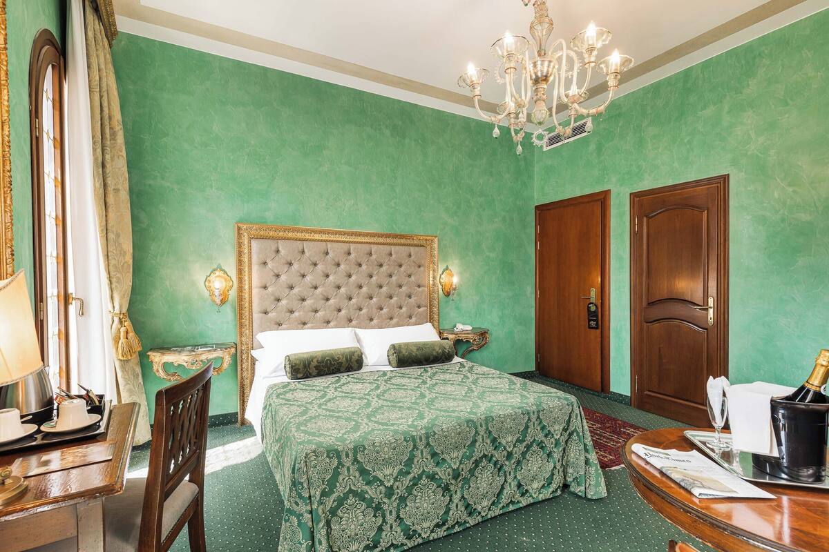 Habitación de hotel con pared verde, cama con cabecera capitonada, escritorio de madera, candelabro y puerta de madera.