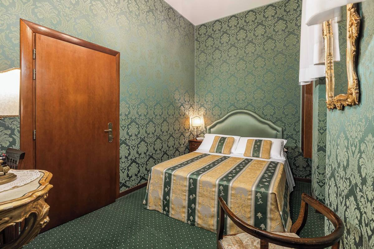 Una habitación con una puerta de madera, una cama con colcha a rayas, una mesita de noche con una lámpara y una silla.