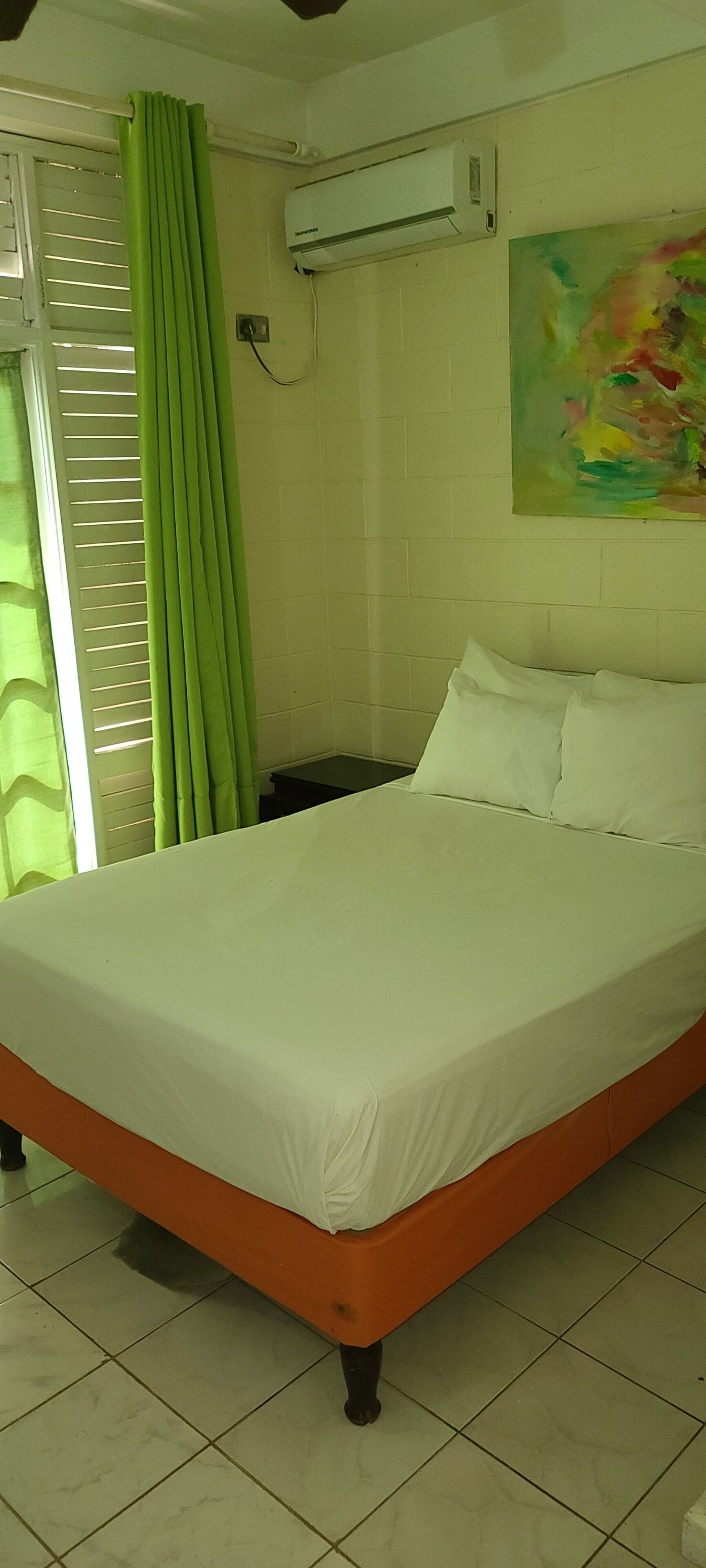 A bed with white sheets in a room with a tiled floor, a green curtain, and a wall-mounted air conditioner.