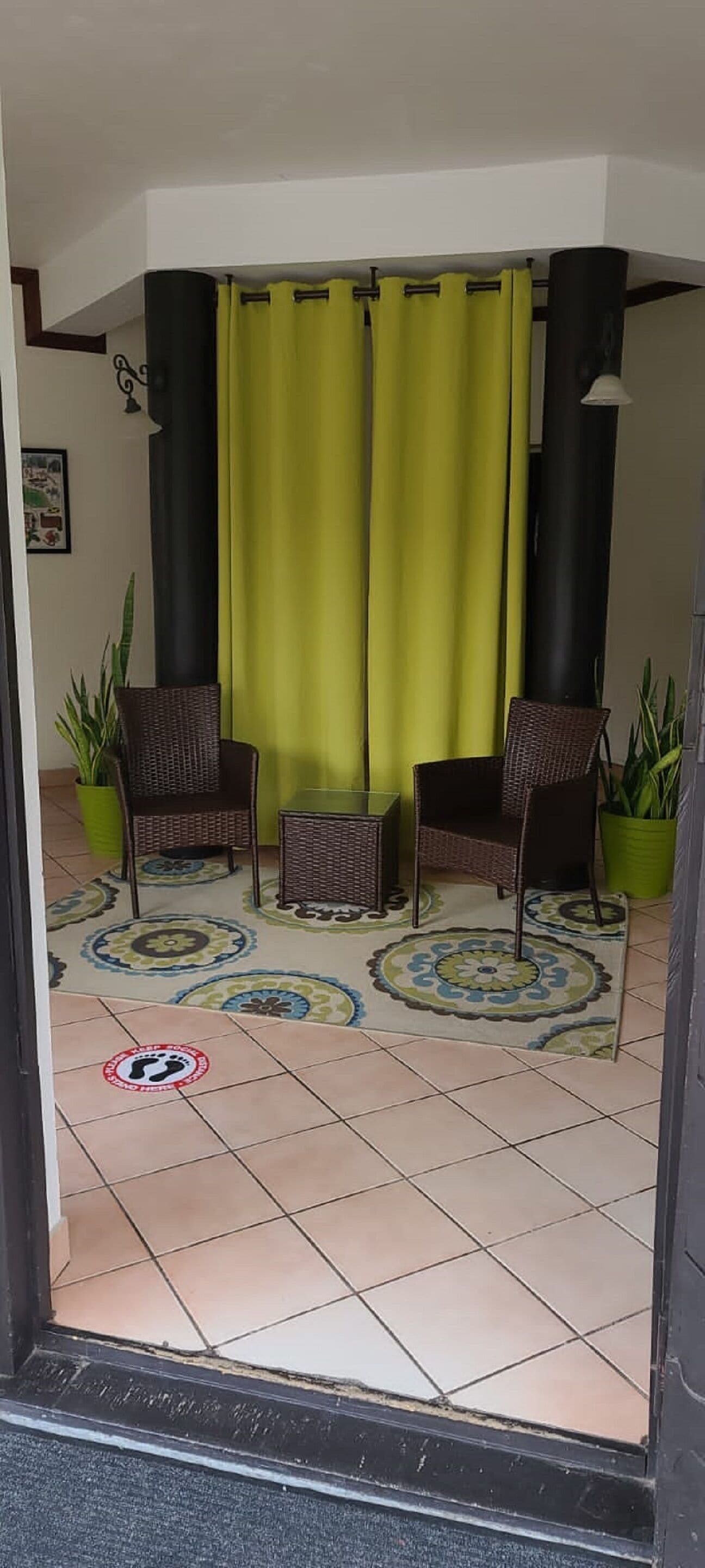 Ein Zimmer mit zwei Korbstühlen, einem kleinen Tisch und einem grünen Vorhang. Es gibt Blumentöpfe und ein Fliesenboden mit einem kreisförmigen Muster.