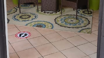 Ein Zimmer mit zwei Korbstühlen, einem kleinen Tisch und einem grünen Vorhang. Es gibt Blumentöpfe und ein Fliesenboden mit einem kreisförmigen Muster.
