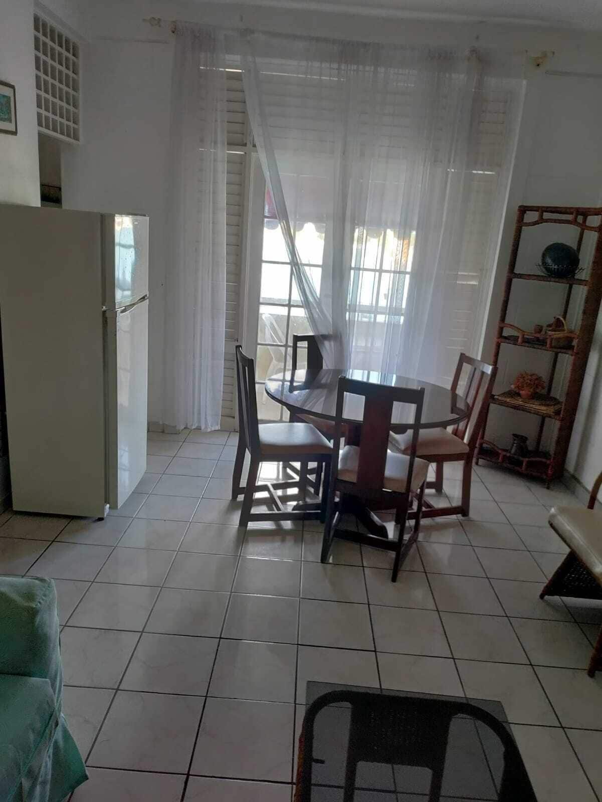 A dining area with a table and chairs, a refrigerator, a window with curtains, and a shelf with items.