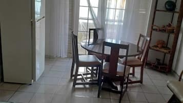 A dining area with a table and chairs, a refrigerator, a window with curtains, and a shelf with items.