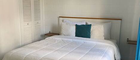 A bedroom with a white bed, a wooden headboard, a white wall, and a window with blue curtains.