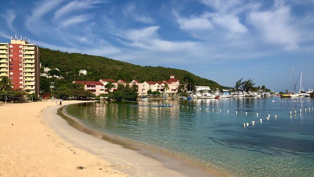 A beach with clear water, a sandy shore, and buildings in the background.