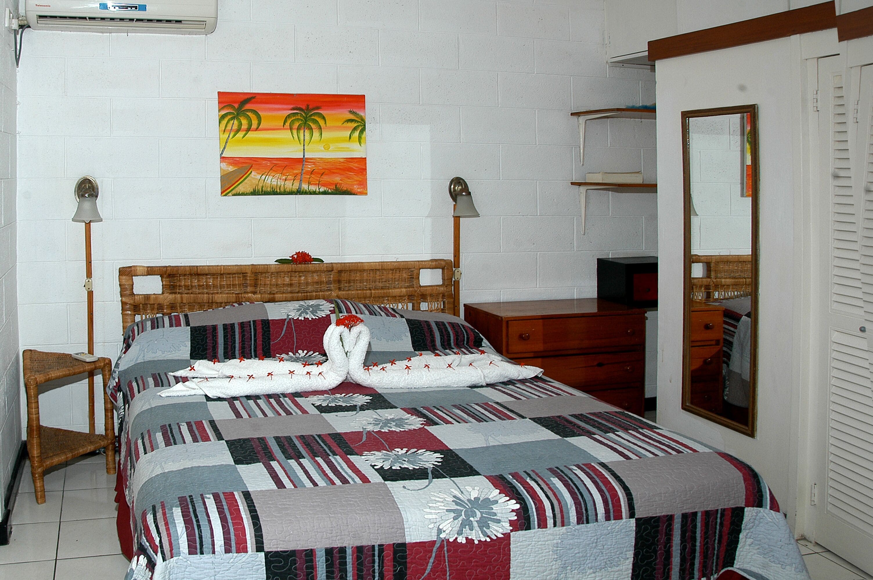Une chambre à coucher comprenant un lit, une table de chevet, une commode, un miroir et un tableau représentant un coucher de soleil.