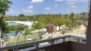 A balcony with white wrought iron chairs and a table, overlooking a pool area with palm trees and a small building.