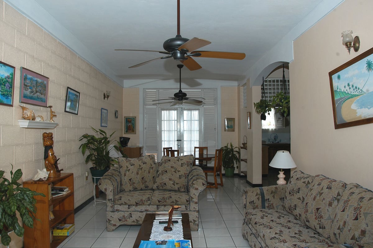 A living room with patterned sofas, a ceiling fan, and a view into the kitchen.