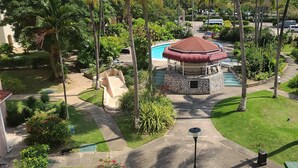 A gazebo with a red roof, surrounded by a well-maintained garden with palm trees and a swimming pool.