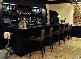 Una zona de bar con una barra alargada, varias estanterías llenas de botellas, taburetes y una lámpara decorativa.