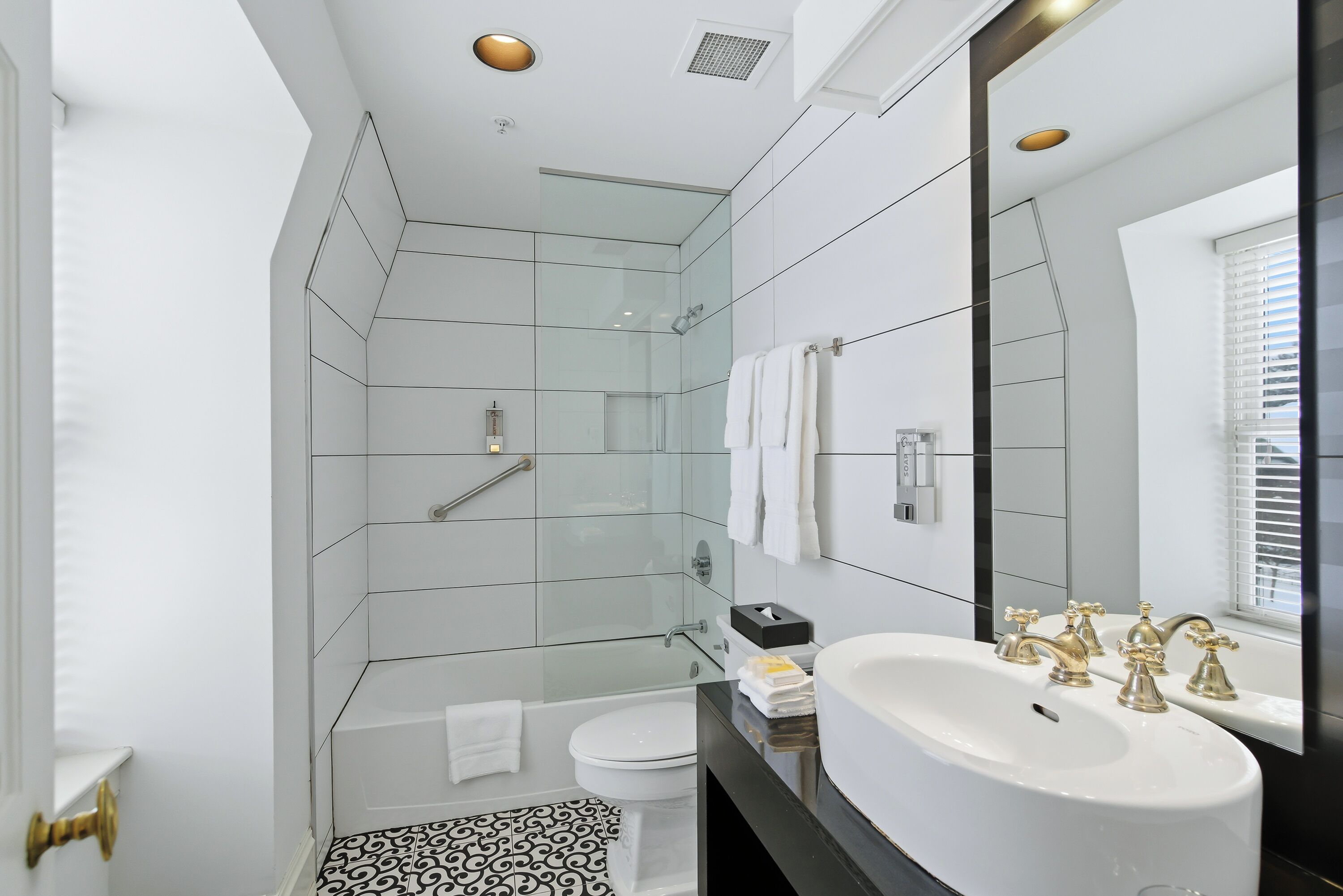 Un baño moderno con cabina de ducha de vidrio, un lavamanos blanco con grifos dorados y un suelo con estampado blanco y negro.