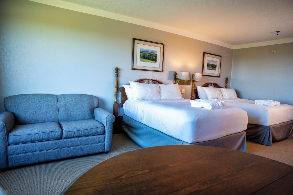 Habitación de hotel con un sofá azul, dos camas con sábanas blancas, una mesa de centro de madera y cuadros enmarcados en la pared.