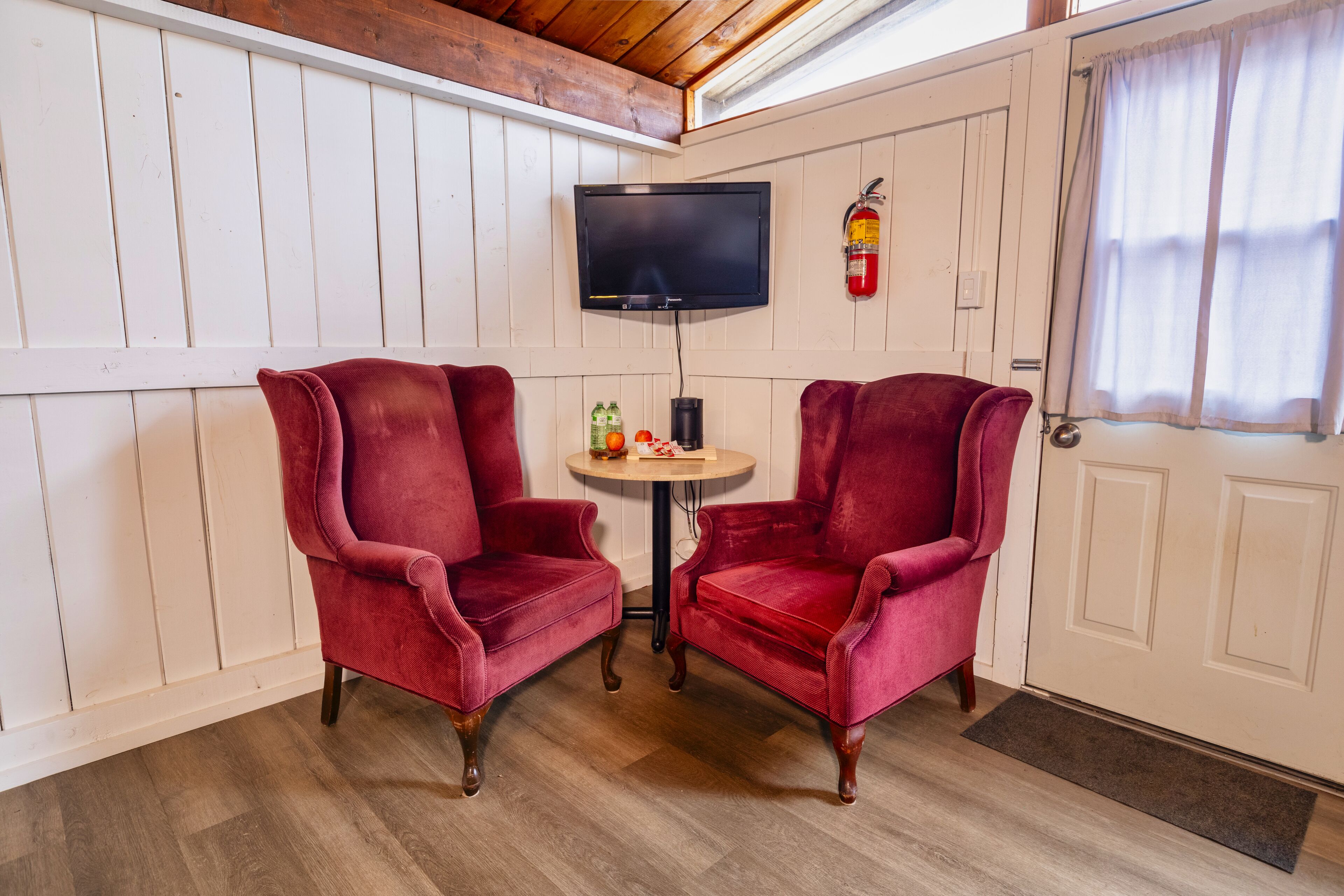 A room with two red velvet chairs, a small round table with a fire extinguisher, and a television mounted on the wall.