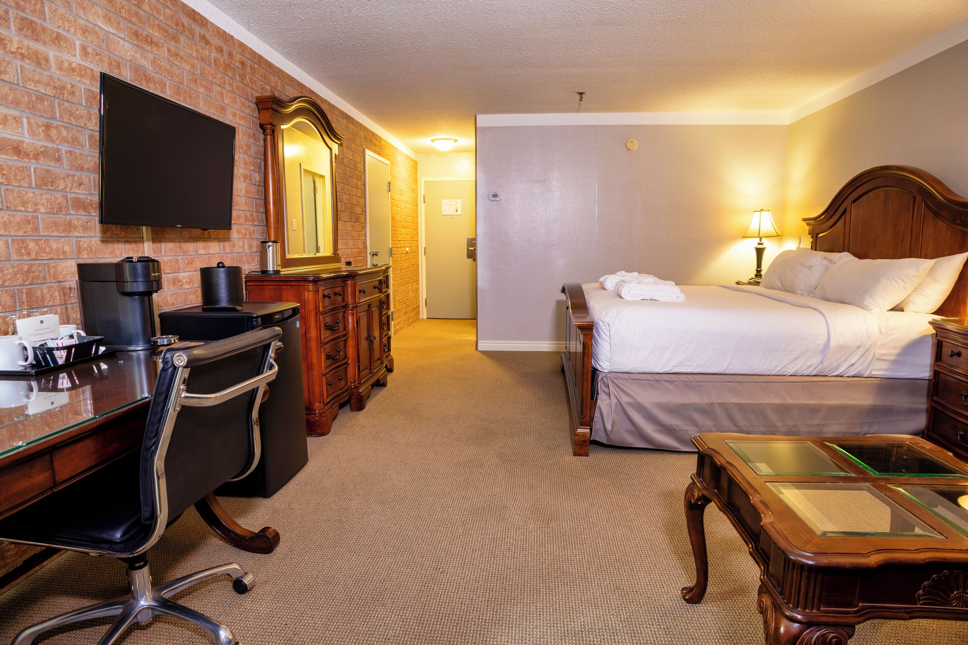 A hotel room with a brick wall, a flat-screen TV, a wooden desk with a chair, a bed with white linens, and a nightstand with a lamp.