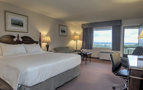 A hotel room with a large bed, a desk, a chair, a sofa, a lamp, a table with wine glasses, and a view of the outdoors.
