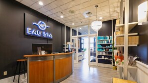 A spa reception area with a wooden reception desk, a computer, and a sign that reads "EAU SPA". There are shelves with products and a chandelier.