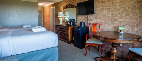 A hotel room with a bed, a dresser, a desk with a chair, a television mounted on the wall, and a brick wall.
