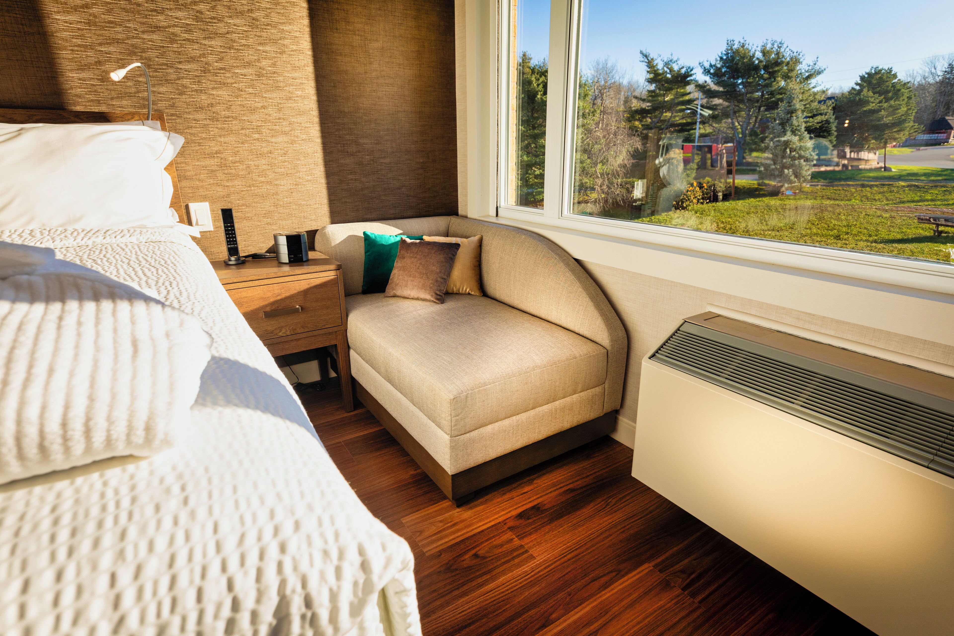 A bedroom with a bed, bedside table, lamp, and a view of a park through a window.