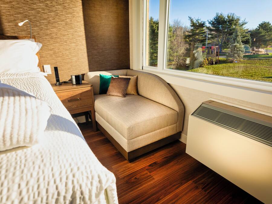 A bedroom with a bed, bedside table, lamp, and a view of a park through a window.