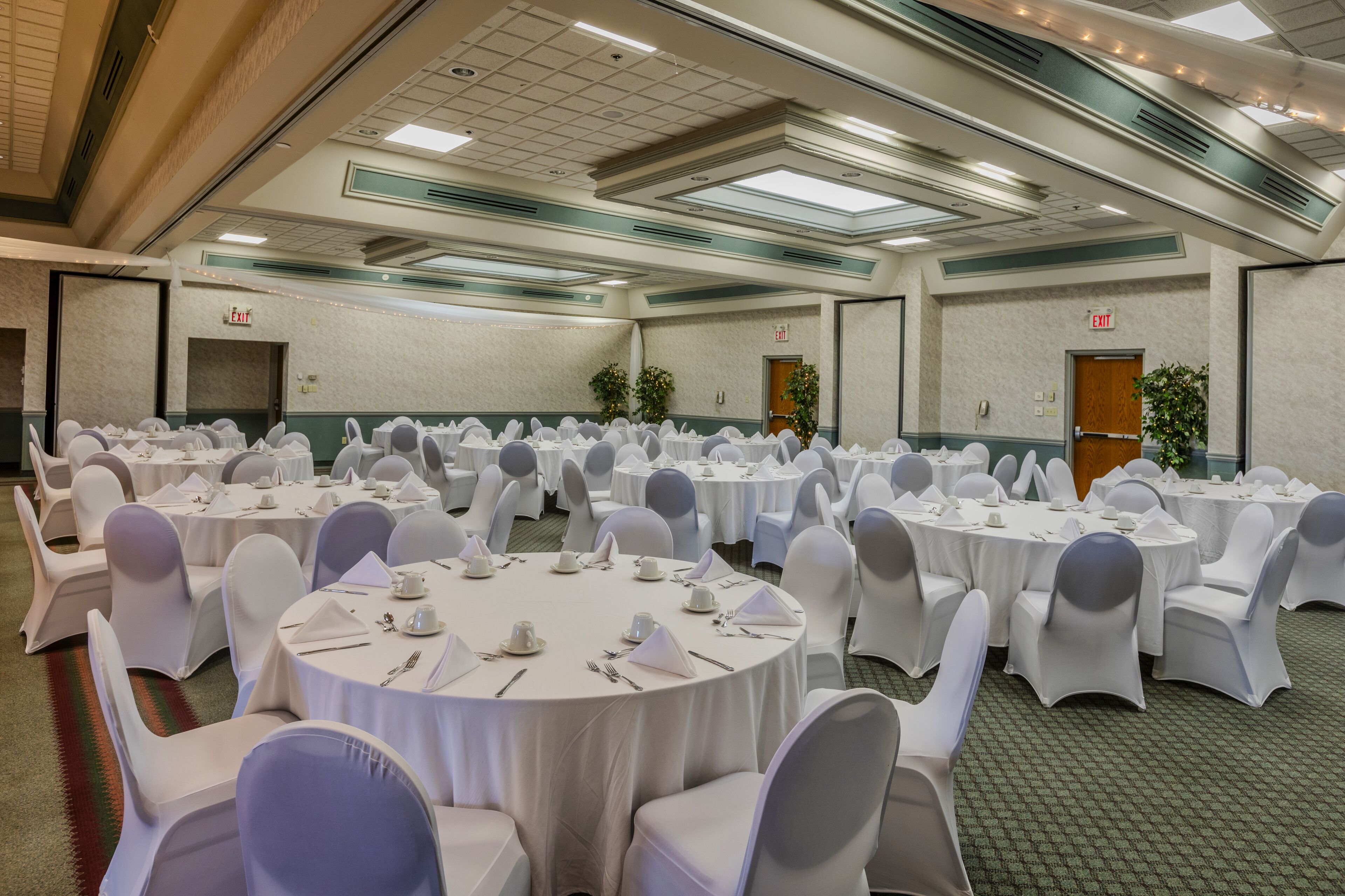 A banquet hall with round tables set for dining, white chairs, and exit signs on the walls.