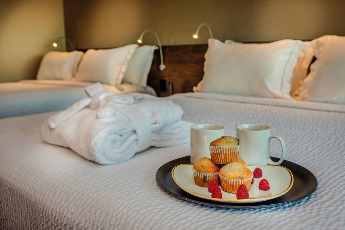 Una cama bien tendida con sábanas blancas, una bandeja con muffins y frambuesas, y dos tazas blancas sobre un plato.