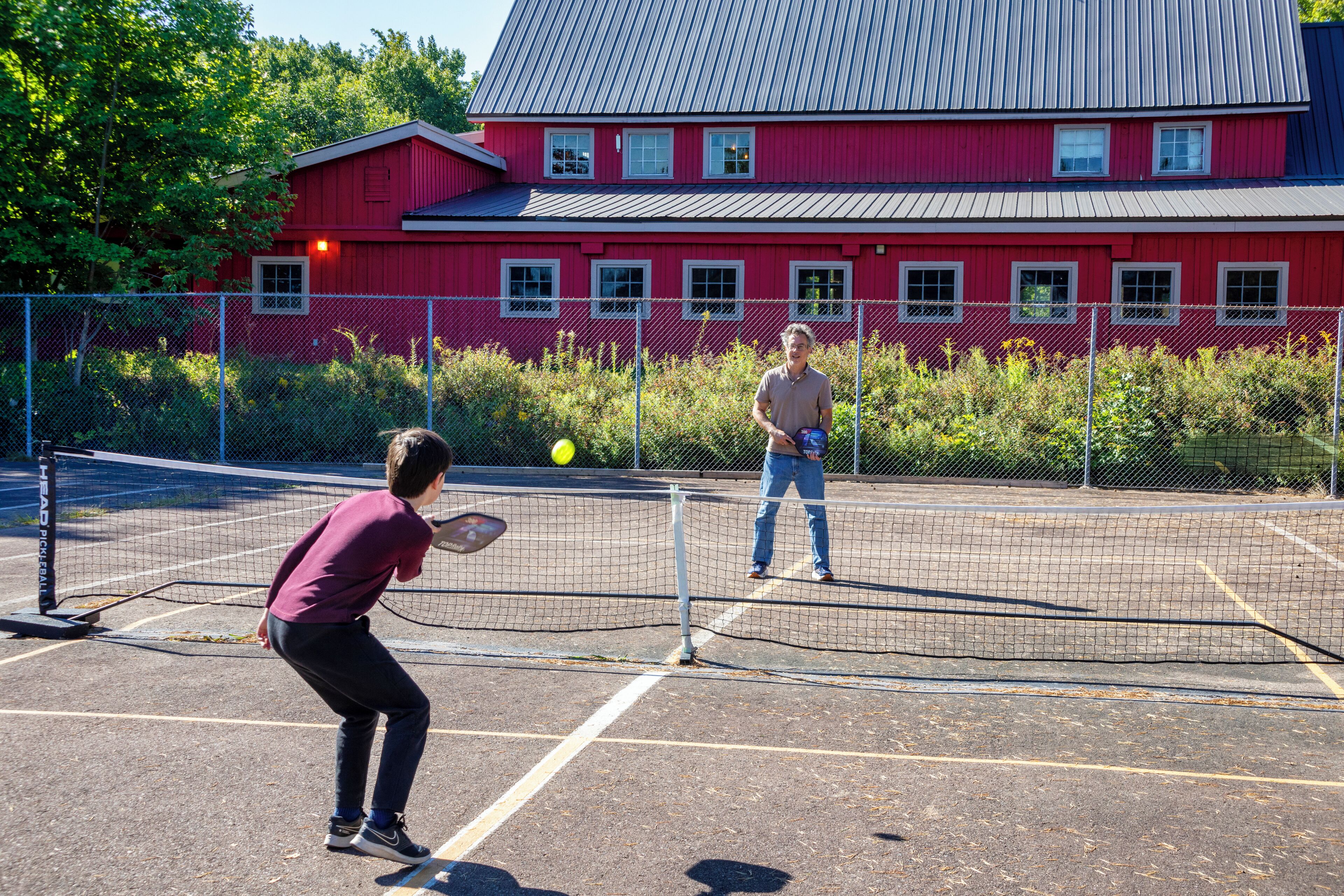 Een tennisbaan met een net, een rood gebouw op de achtergrond en een persoon die tennist.