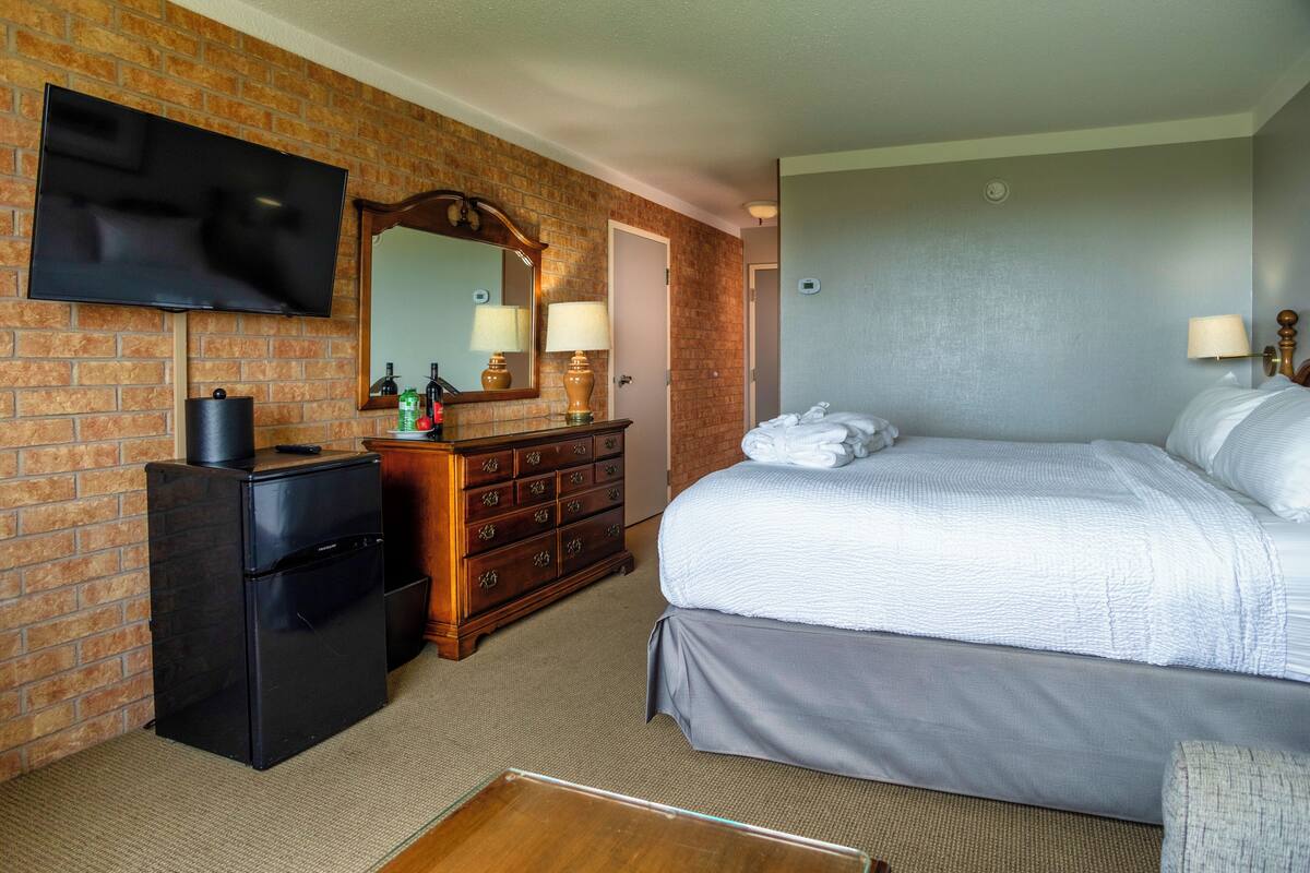 Habitación de hotel con pared de ladrillo, televisor de pantalla plana, cómoda de madera, cama con sábanas blancas y un refrigerador pequeño.