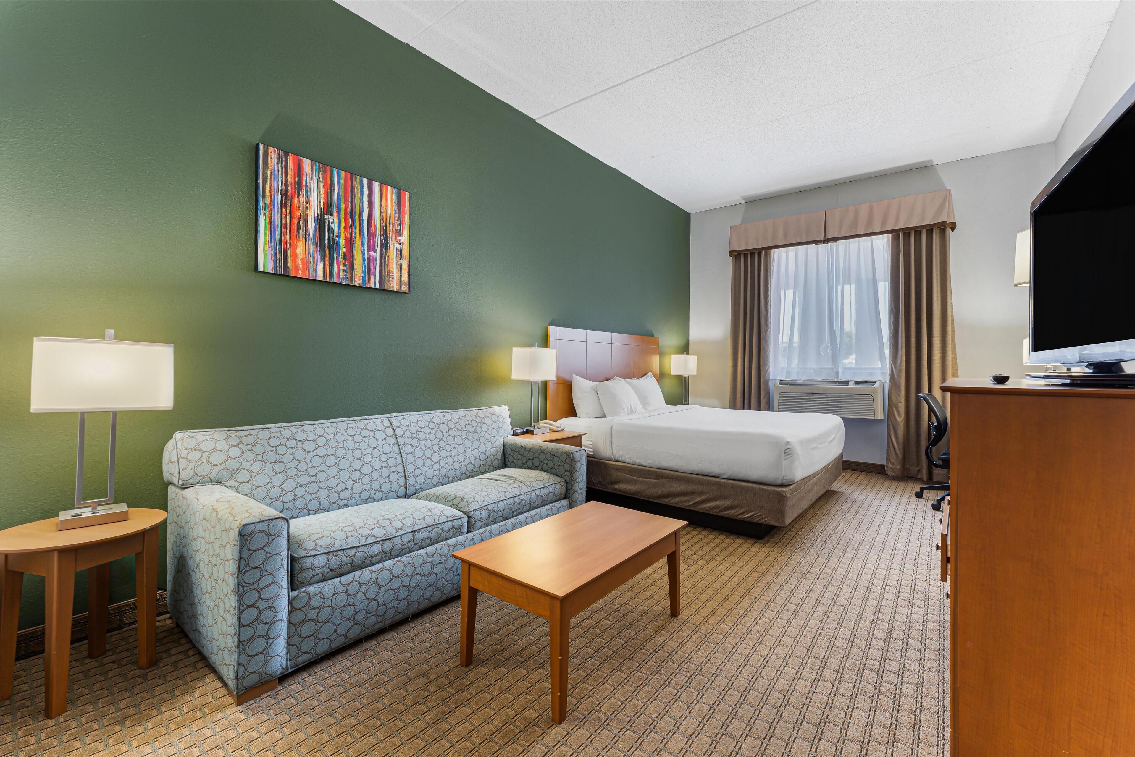 Una habitación de hotel con cama, sofá, mesa de centro, televisor y un cuadro en la pared.