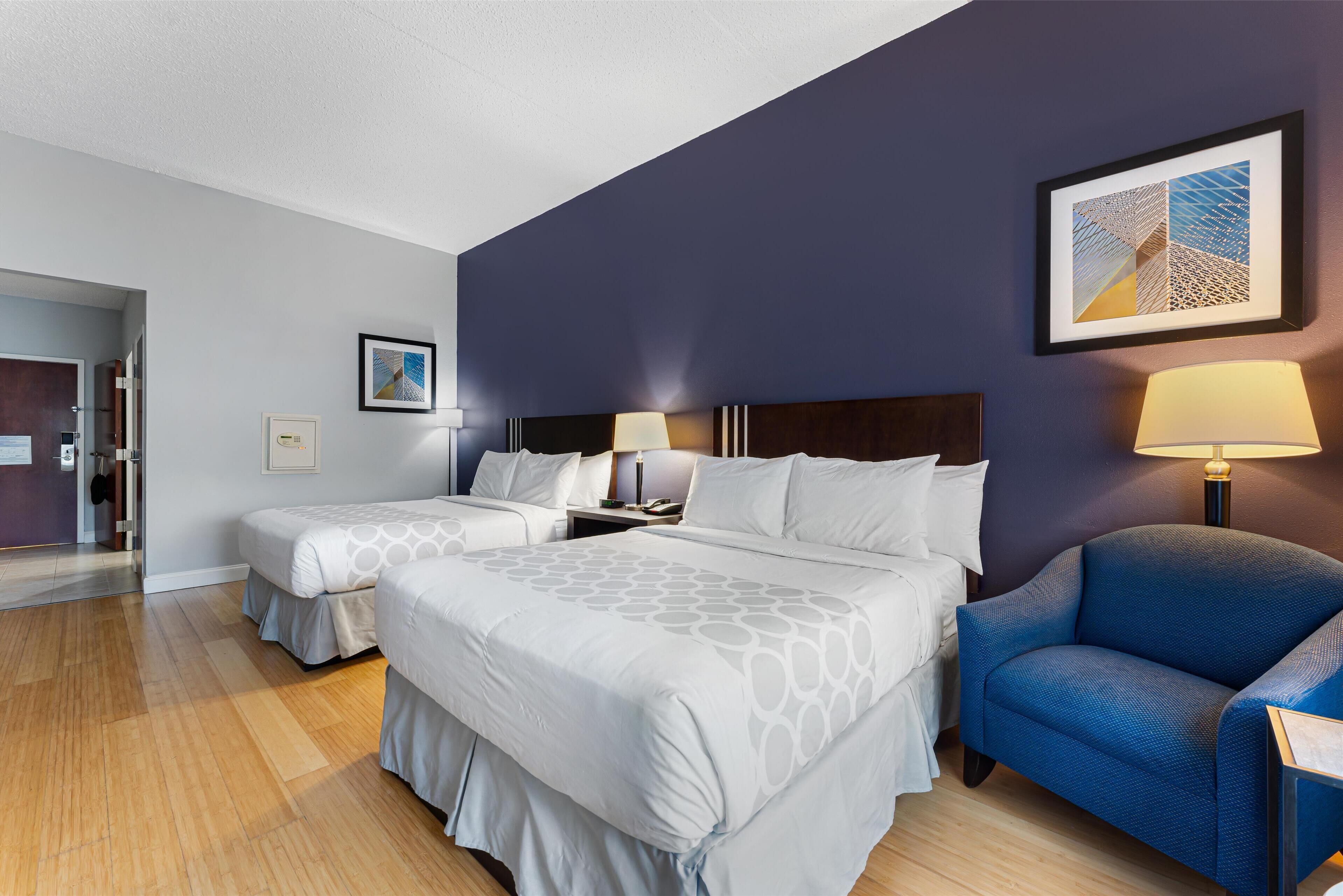 A hotel room with two beds, a blue armchair, and a framed artwork on the wall.
