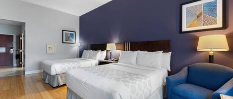 A hotel room with two beds, a blue armchair, and a framed artwork on the wall.