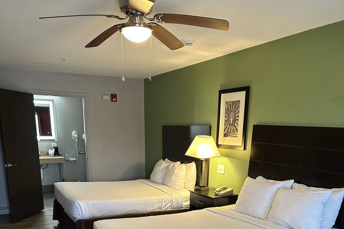 A hotel room with two beds, a ceiling fan, a nightstand with a lamp, and a bathroom visible through an open door.
