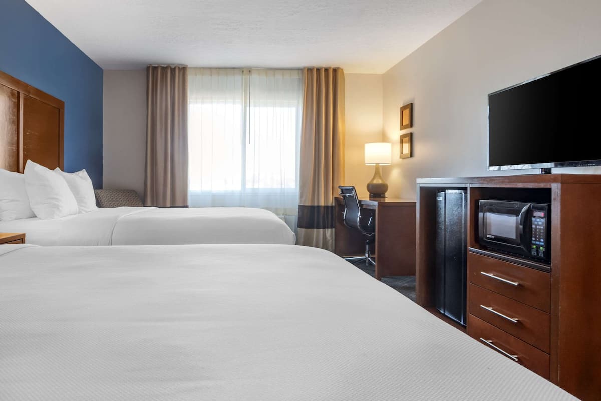 A hotel room with two beds, a desk with a chair, a flat-screen TV, a microwave, and a window with curtains.