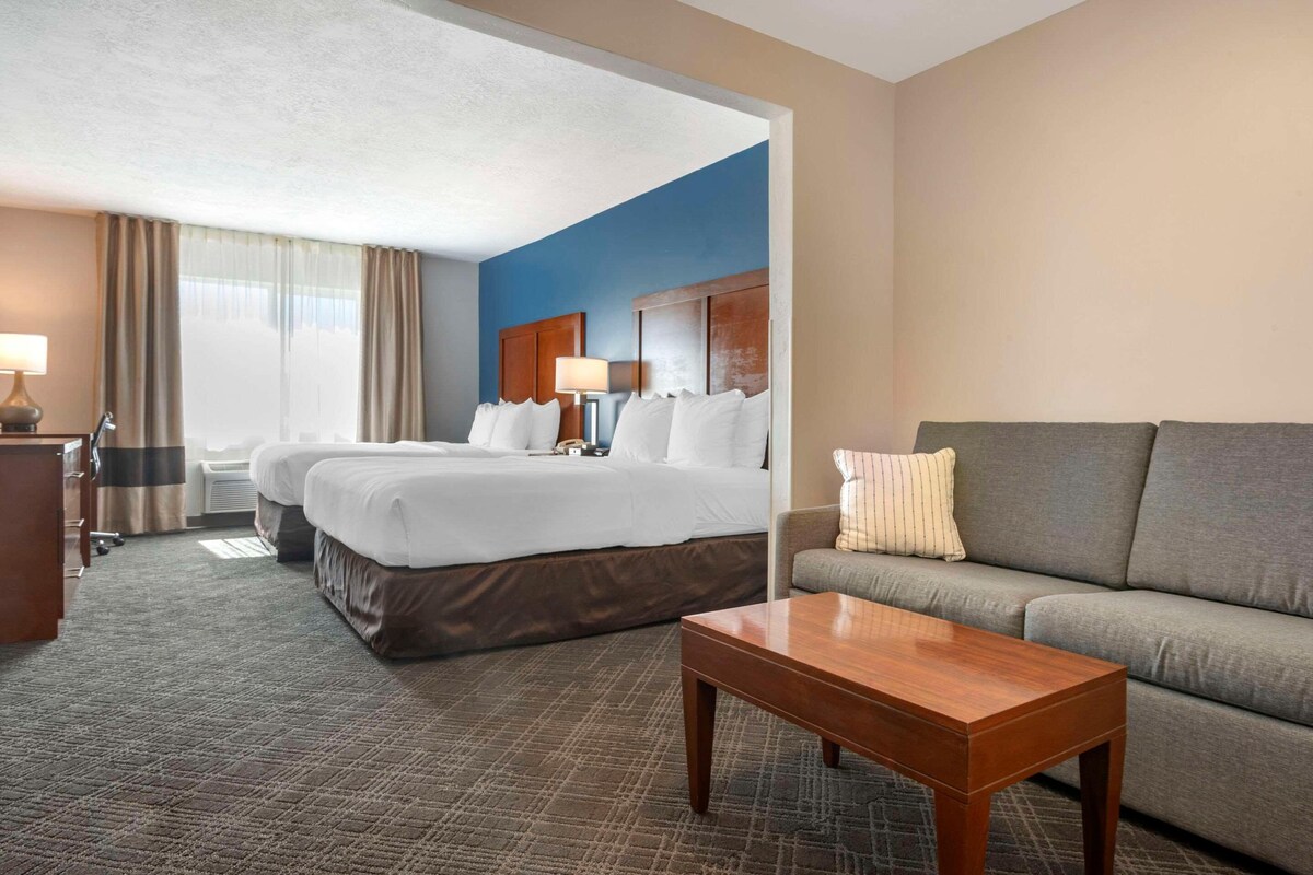 A hotel room with two beds, a sofa, a wooden table, and a desk with a lamp.