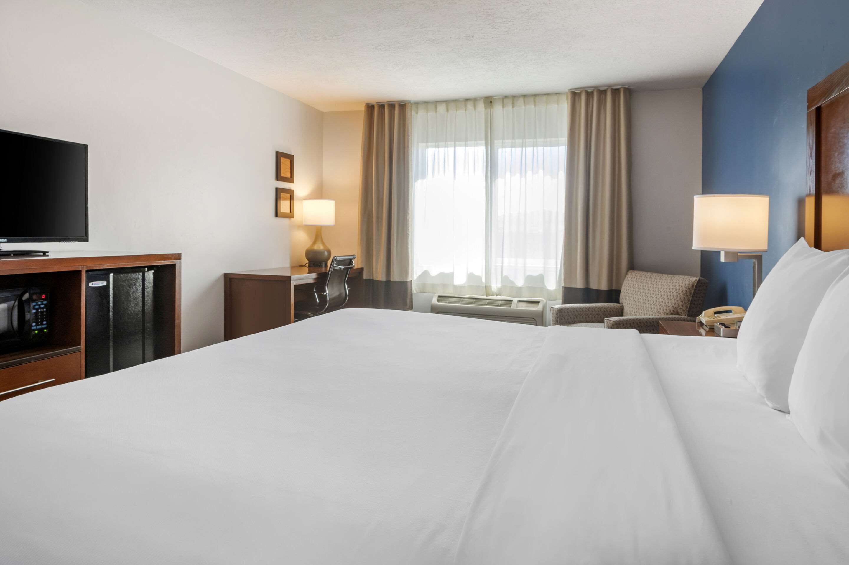 A hotel room with a large bed, a flat-screen TV, a desk with a lamp, a chair, and a window with curtains.