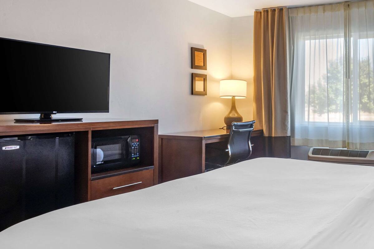 A hotel room with a flat-screen TV, a desk with a lamp, a chair, a bed, and a minibar.