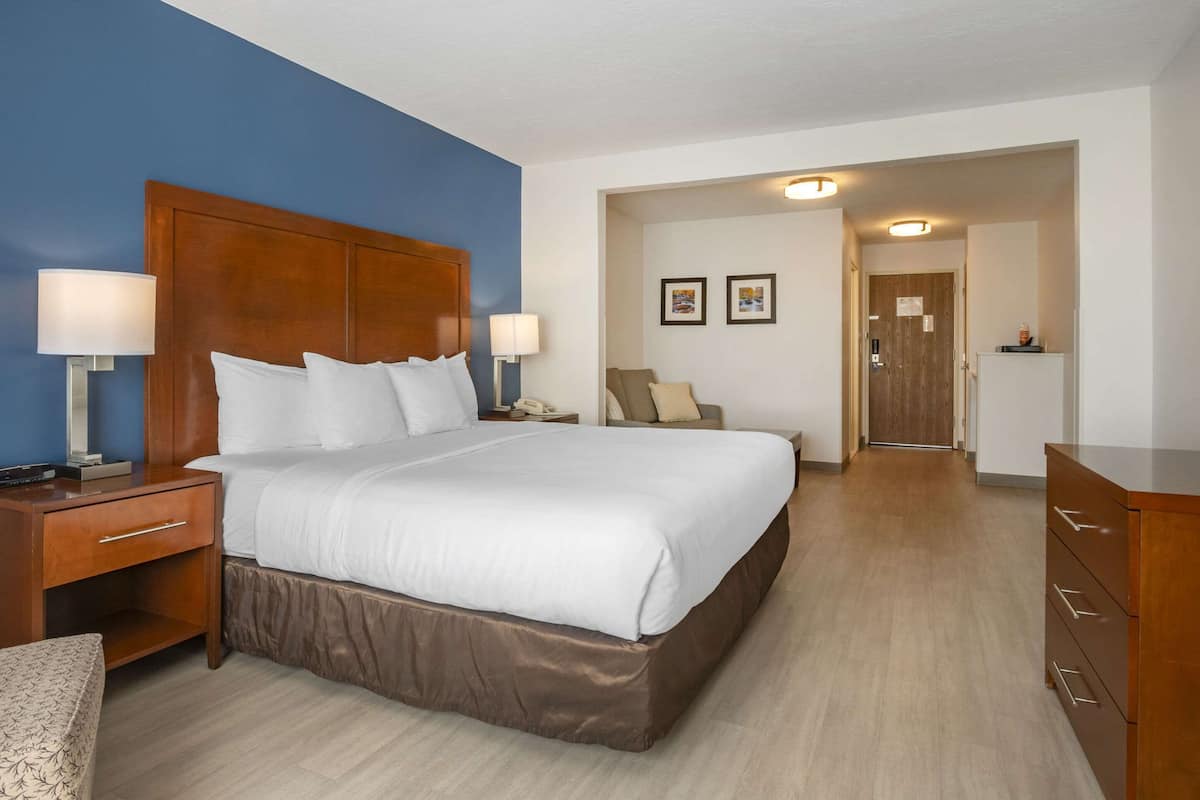 A hotel room with a large bed, wooden headboard, two bedside tables with lamps, a nightstand, a dresser, and a door leading to another room.