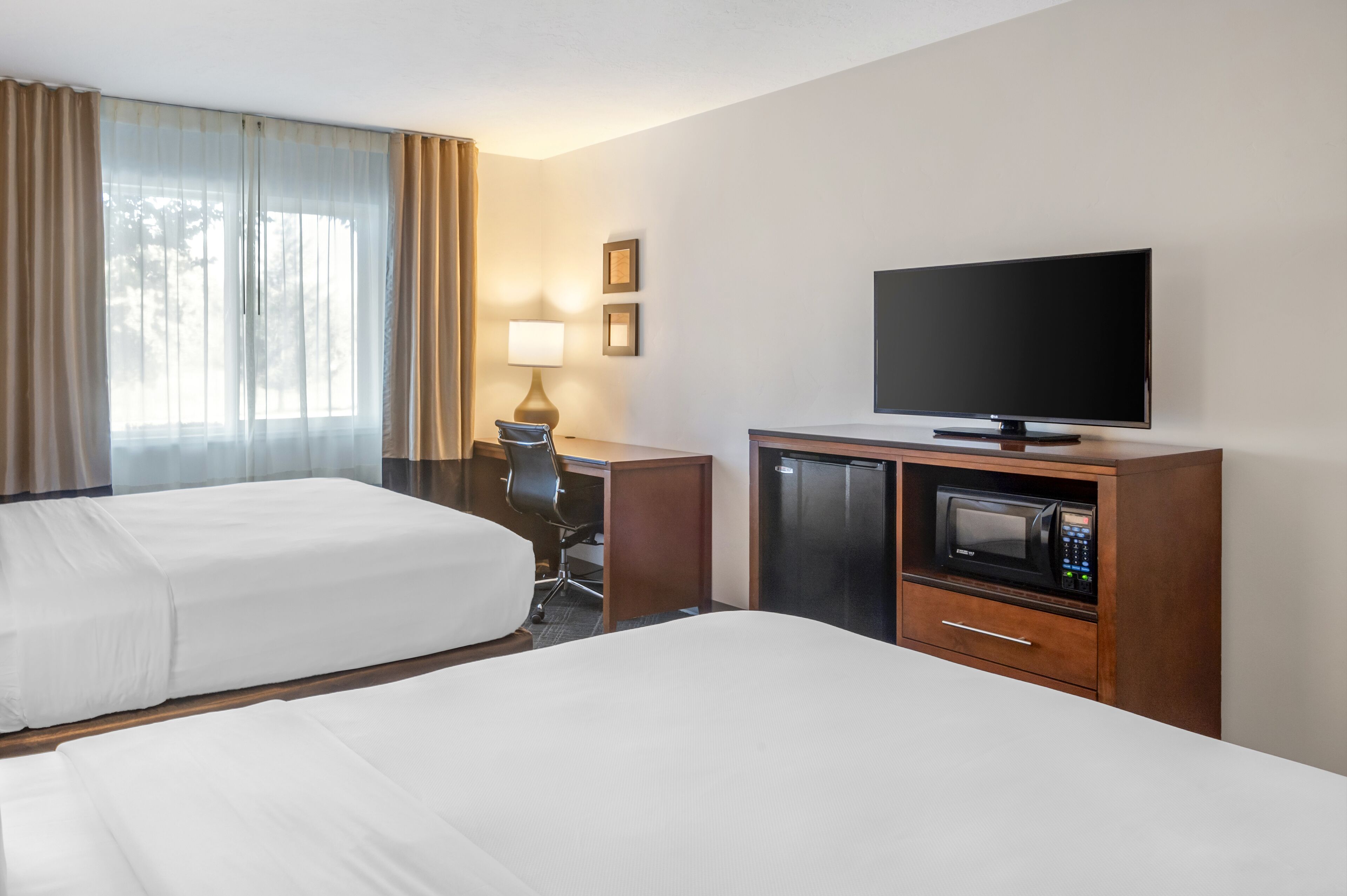 A hotel room with two beds, a desk with a lamp, a television, a refrigerator, and a microwave.