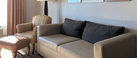 A hotel room with a beige sofa, a striped armchair, a small ottoman, a floor lamp, and framed artwork on the wall.