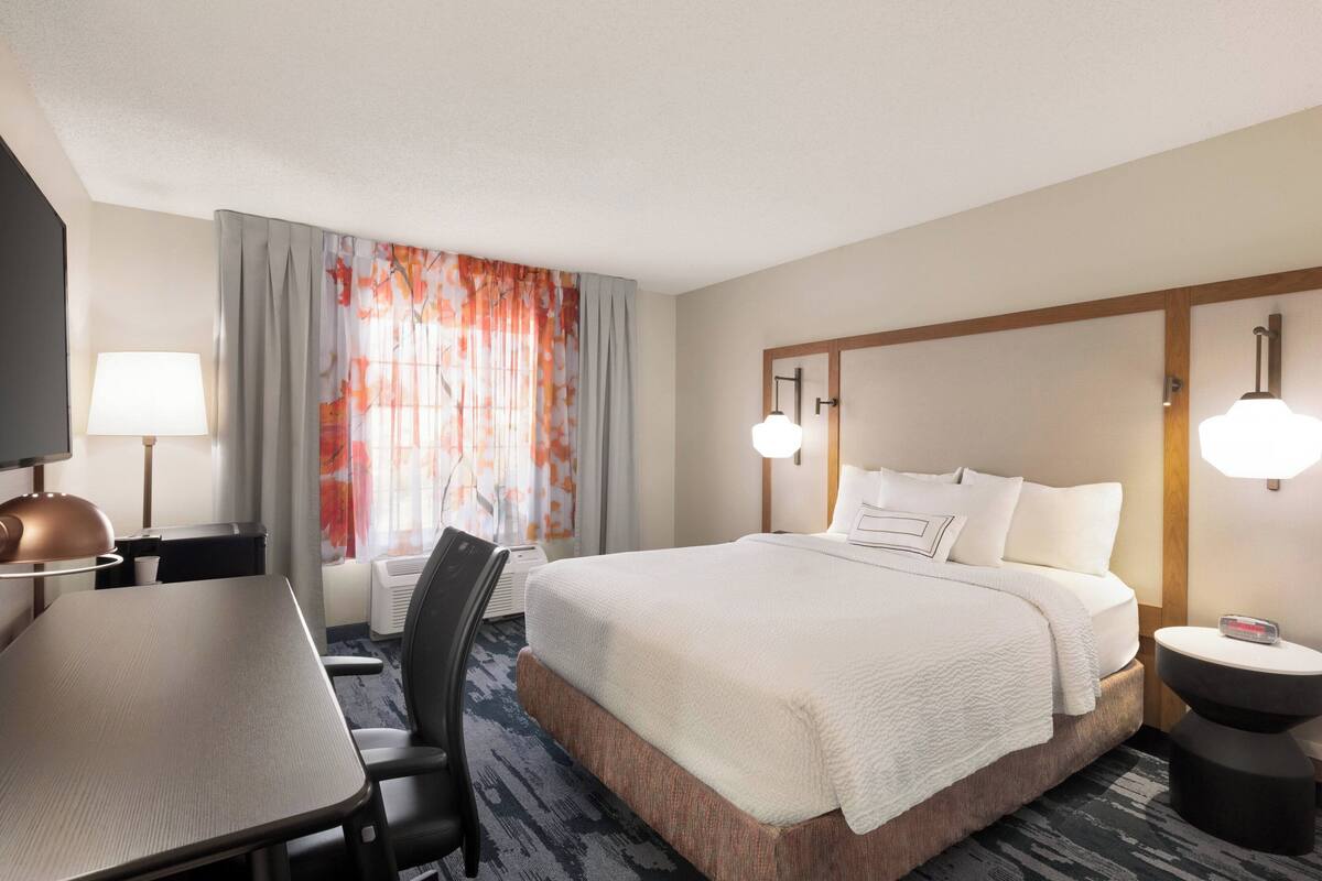 Ein Hotelzimmer mit Bett, Schreibtisch, Stuhl, Lampe und einem fenster mit Vorhängen.
