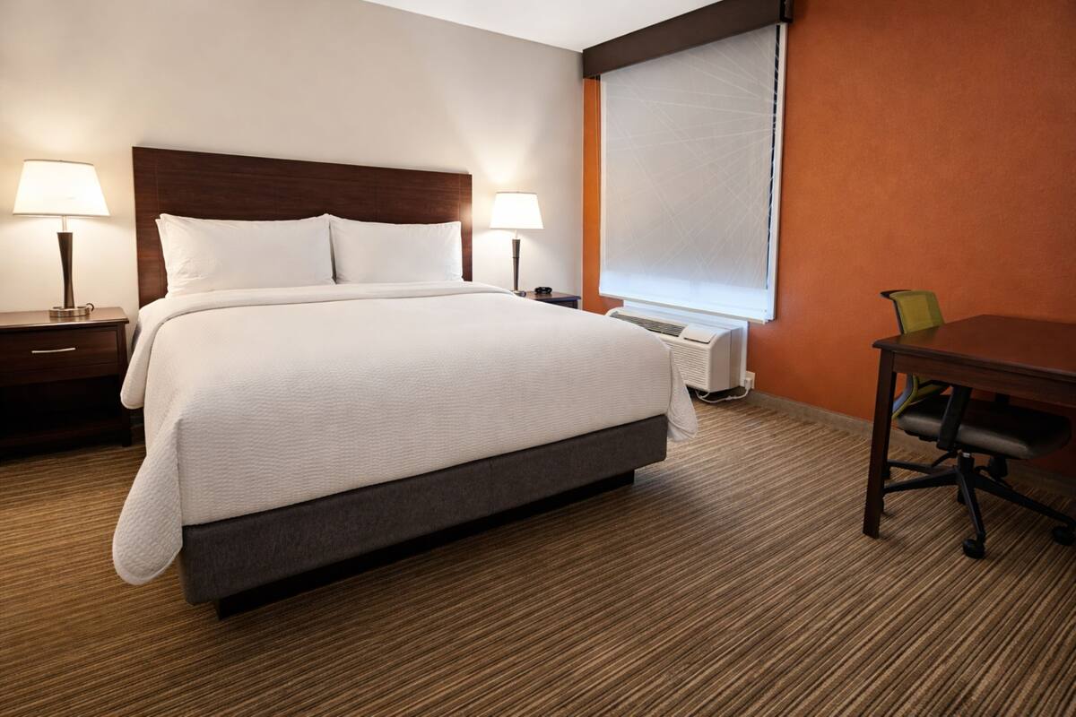 Una habitación de hotel con cama, dos lámparas de noche, un escritorio con silla y una ventana con persianas.