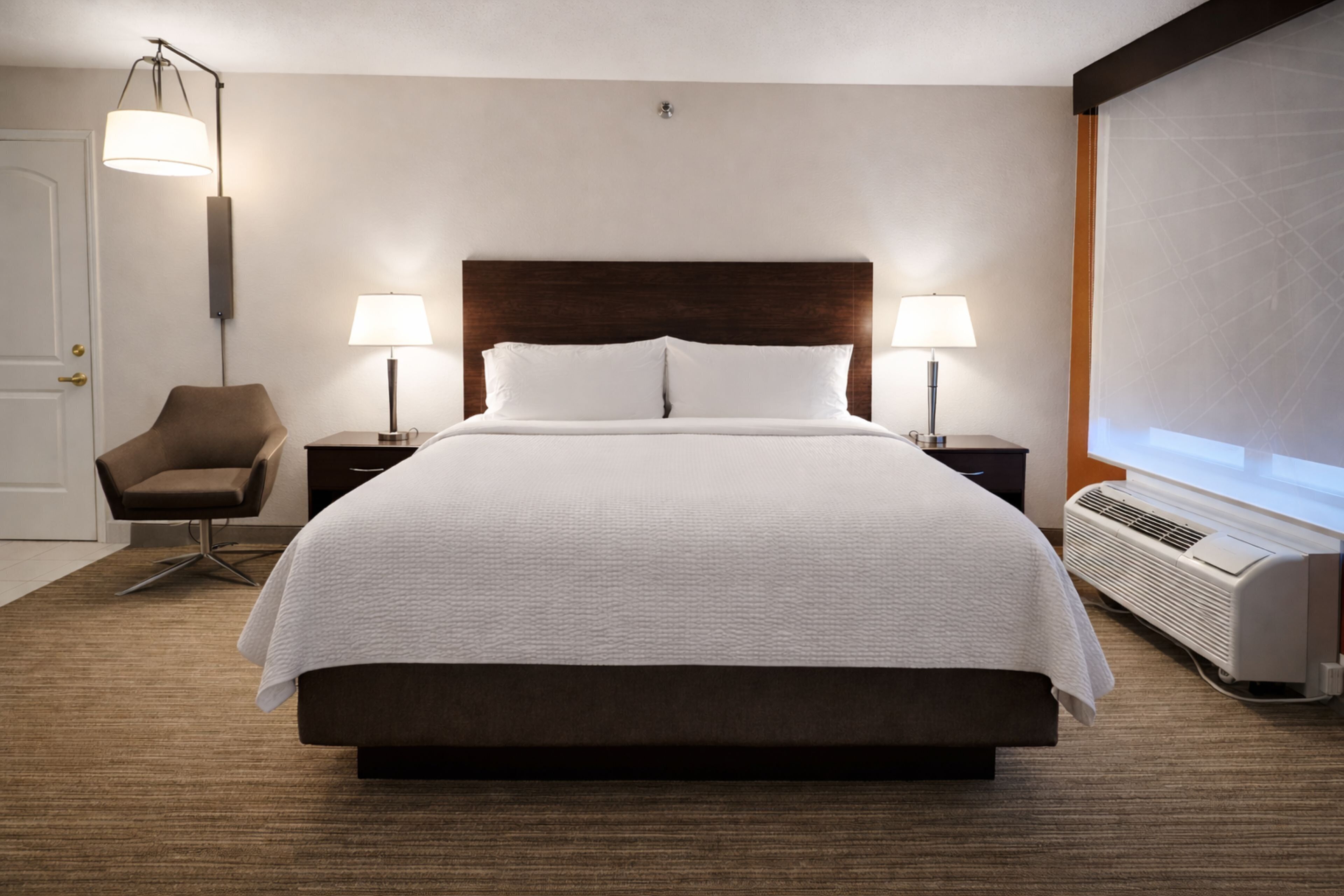 A hotel room with a large bed, two bedside tables with lamps, a chair, a heater, and a door.