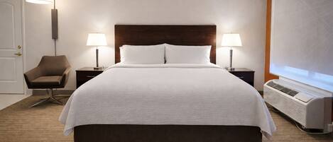 A hotel room with a large bed, two bedside tables with lamps, a chair, a heater, and a door.