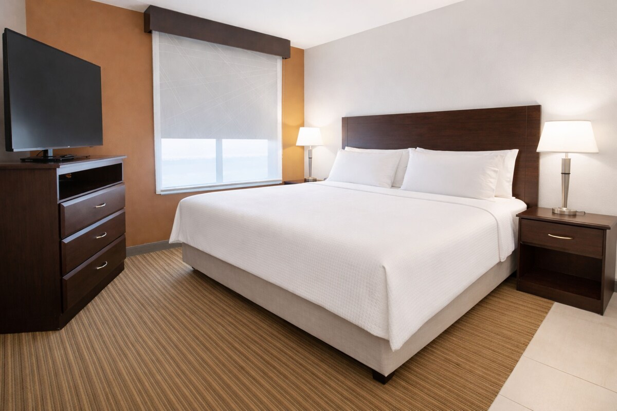 Una habitación de hotel con una cama grande, dos mesitas de noche con lámparas, un televisor de pantalla plana en una cómoda y una ventana con persianas.