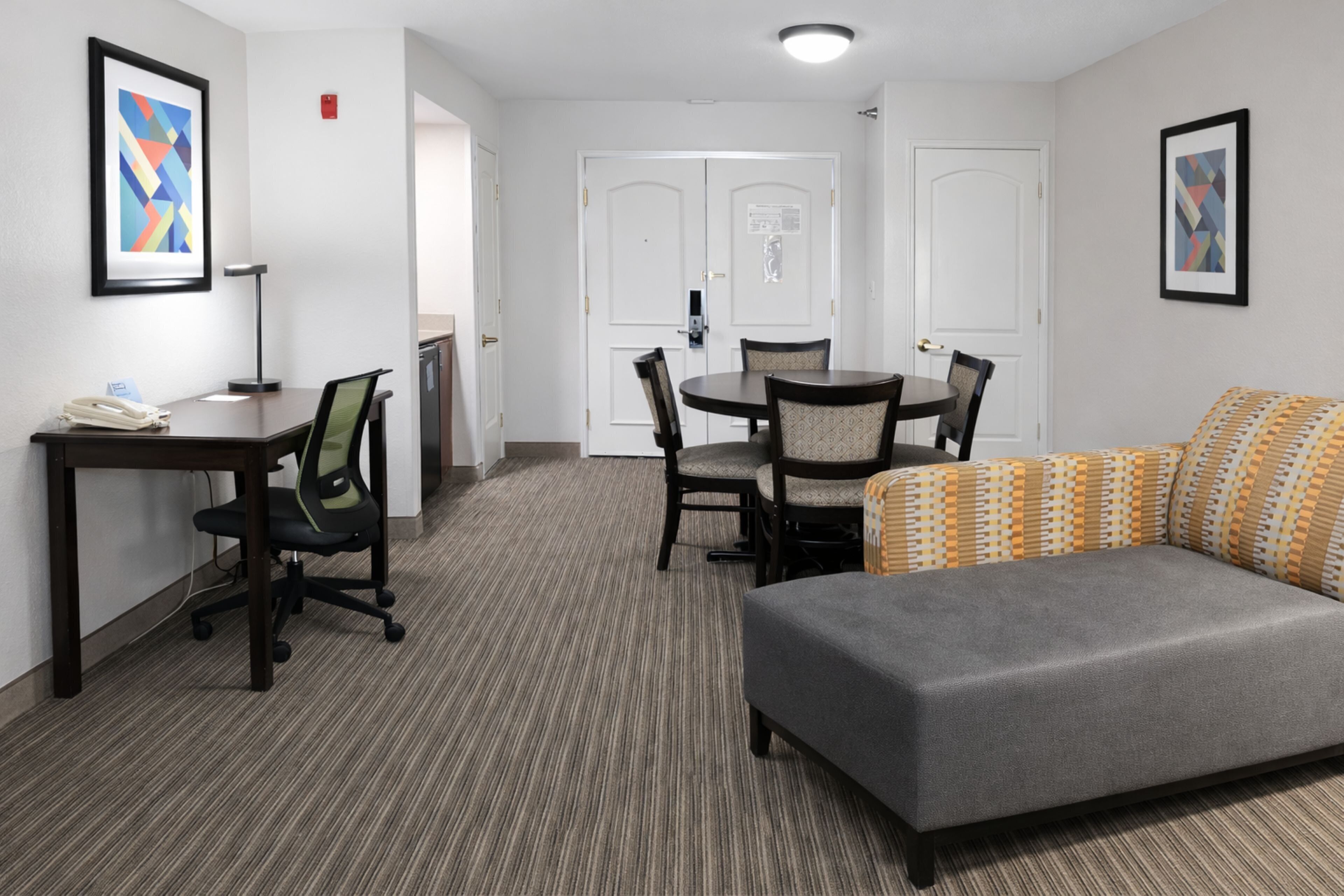 A hotel room with a gray sofa, a small table with a lamp, a chair, and a door with a notice.