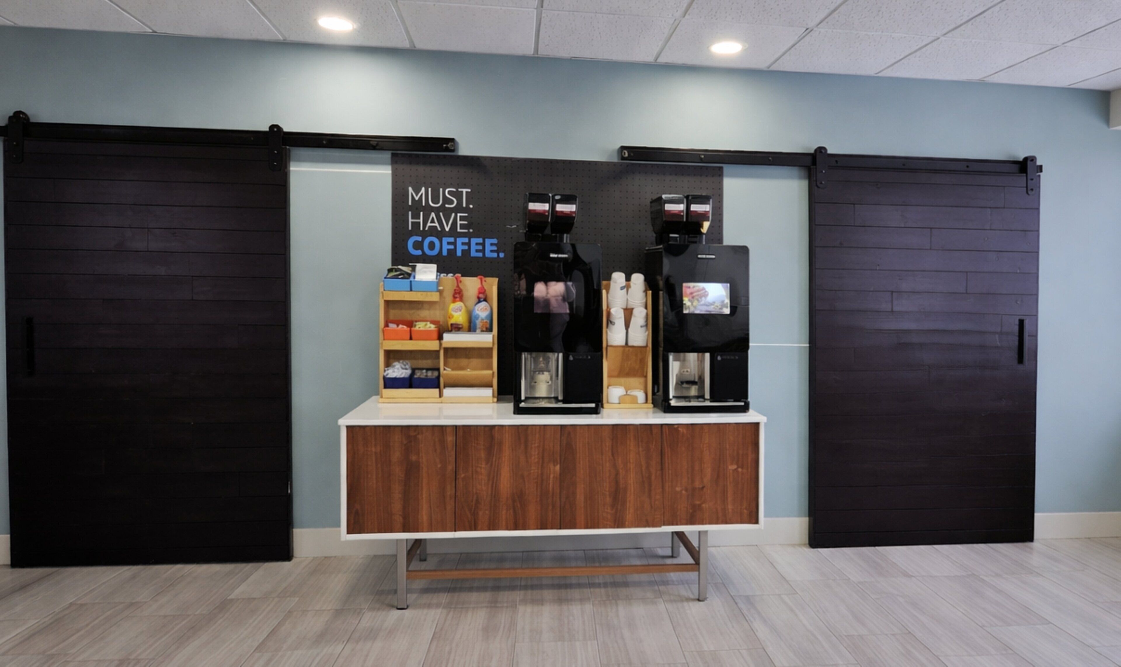 Een koffiehoek met twee espresso machines, een bord waarop staat 'COFFEE IS ESSENTIEEL' en een plank met diverse koffieproducten.
